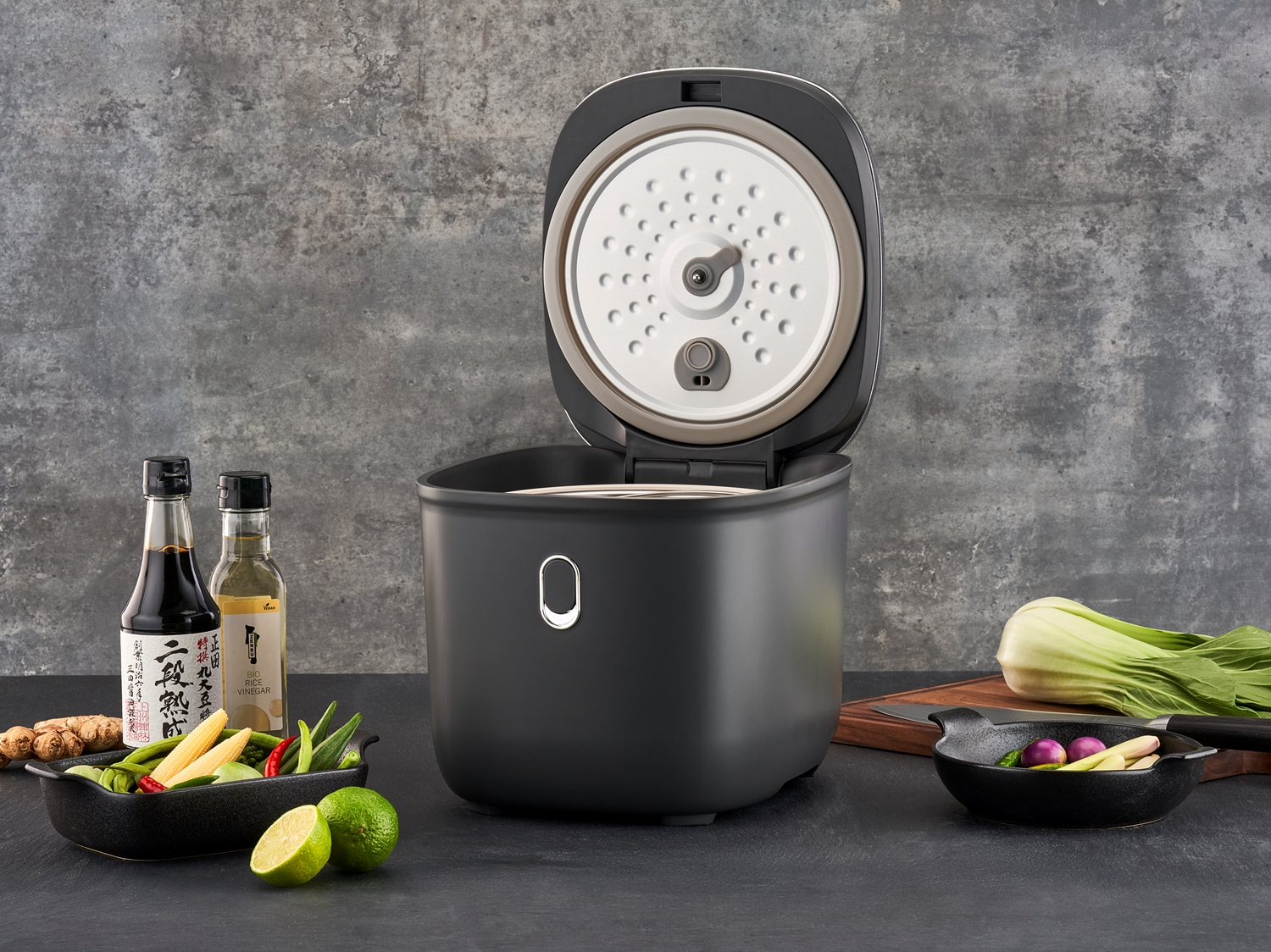 Cuiseur à riz ouvert au design moderne, entouré d’ingrédients frais et d’assaisonnements asiatiques.
